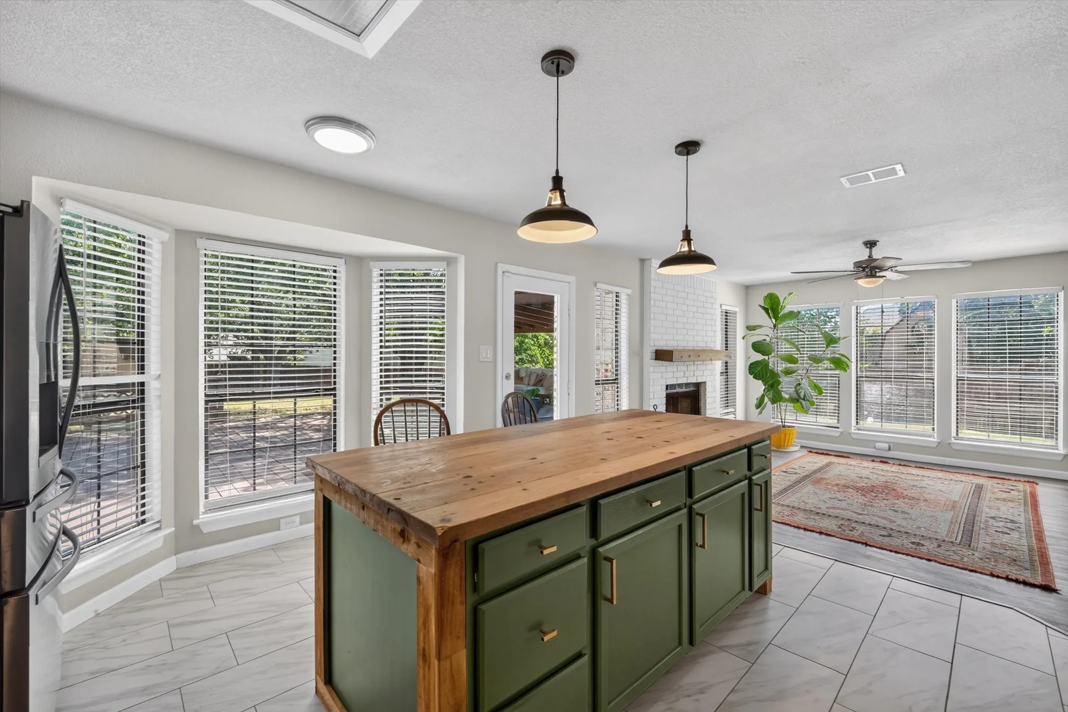 Kitchen island with wood-finish countertop and green cabinetry, bay window, white brick-finish fireplace, and ceiling fan