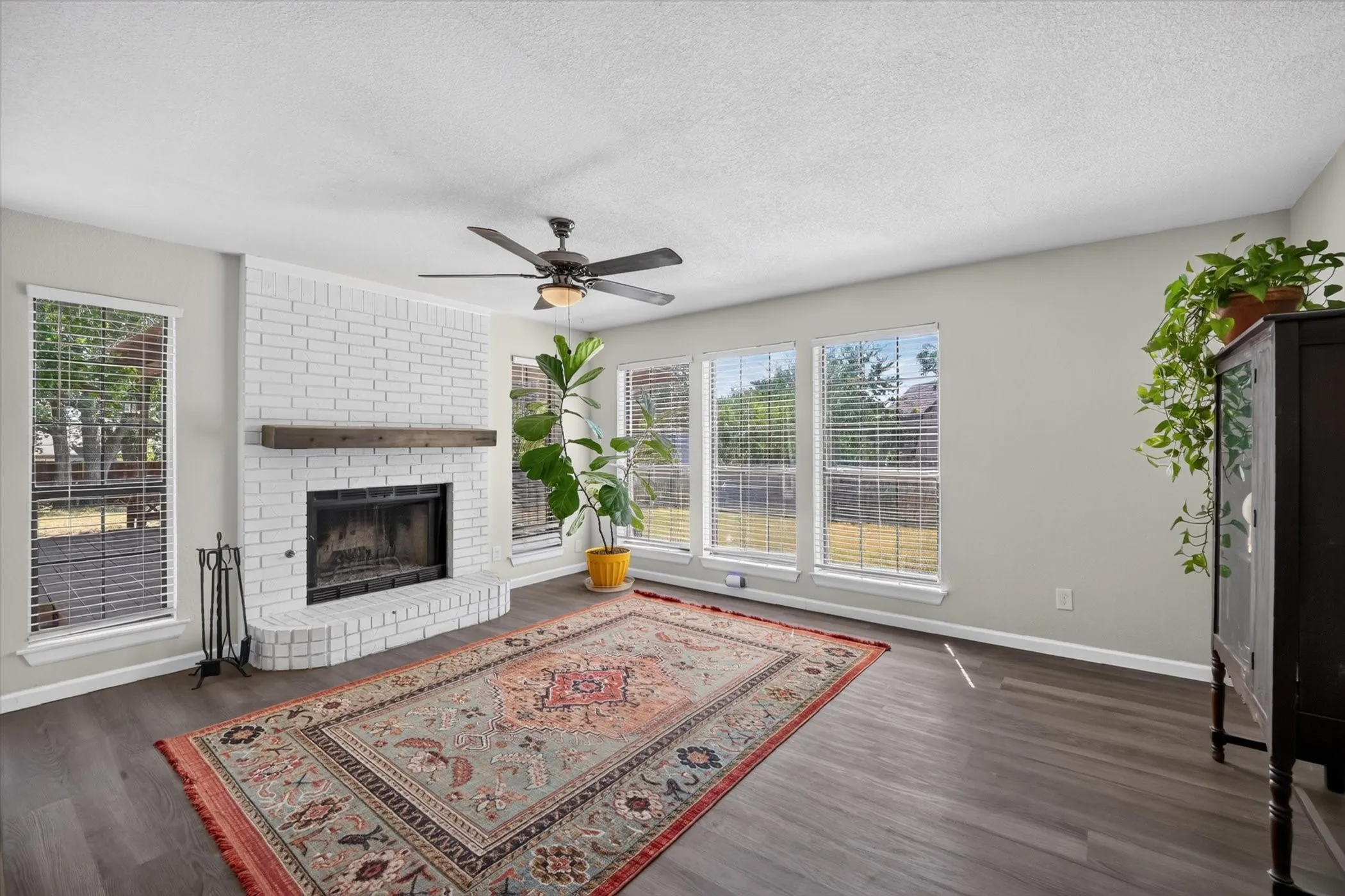 White-painted brick fireplace with a wood mantle, ceiling fan, dark wood-finish flooring, and multiple windows