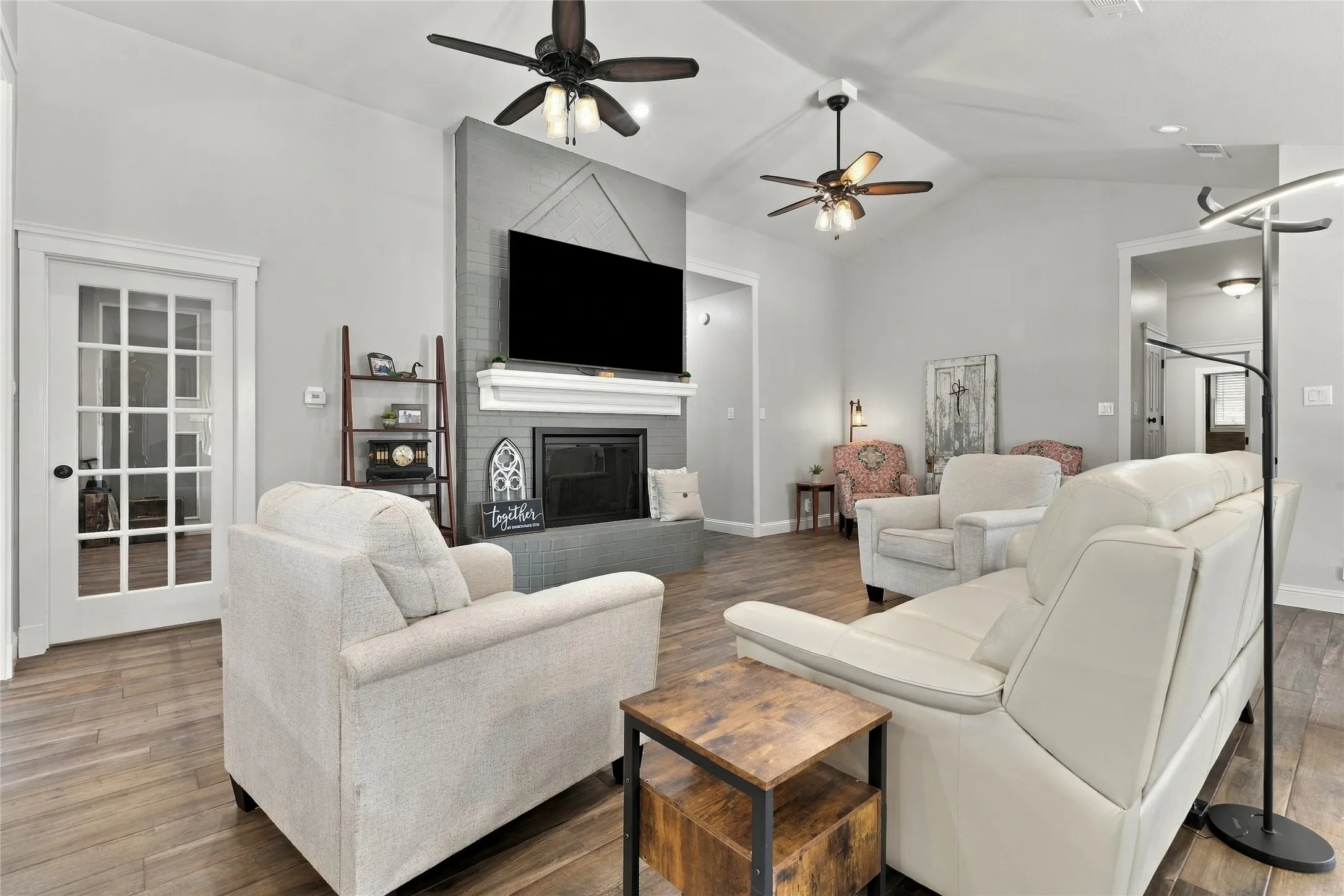 Vaulted ceiling with dual ceiling fans, wood-finish flooring, white French doors, and a fireplace with a mantel