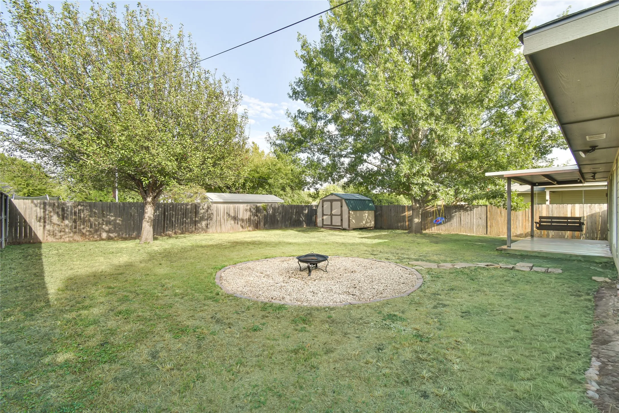 Expansive backyard featuring a privacy fence, mature trees, a fire pit area with gravel, and a covered patio with an outdoor swing