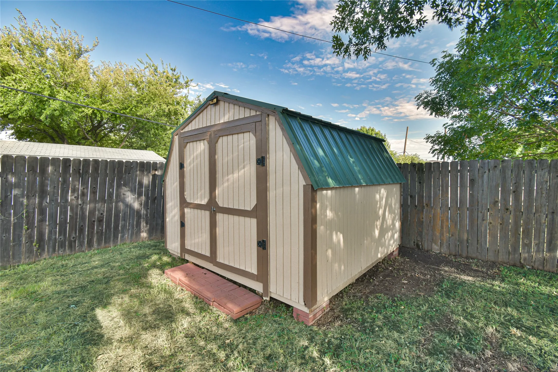 Detached storage shed with a gambrel-style green metal roof and dual wood doors, set within a fenced yard