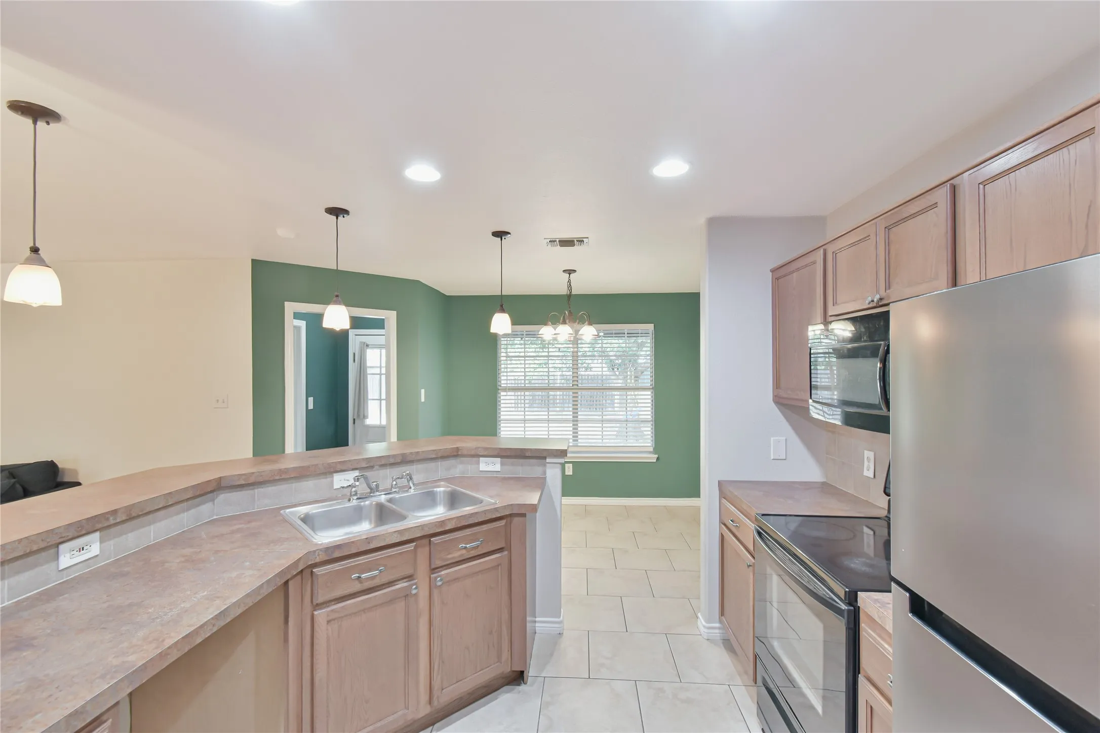 Kitchen featuring a stainless steel double sink, light wood-finish cabinetry, stainless steel refrigerator, built-in microwave, and tile flooring