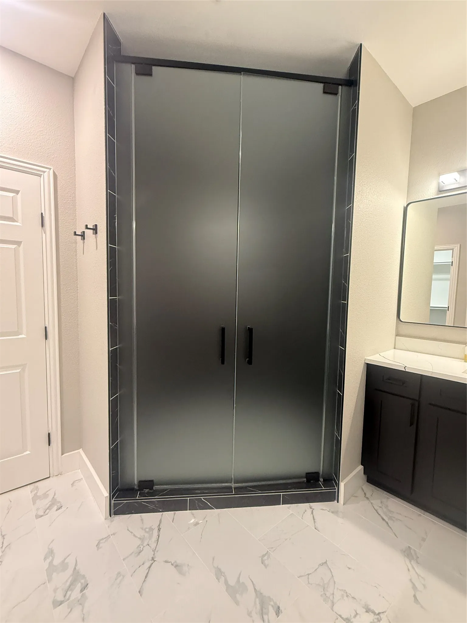 Frosted glass shower enclosure with matte black hardware, black subway tile shower surround, dark wood-finish vanity with white countertop, and large format marble-look floor tiles