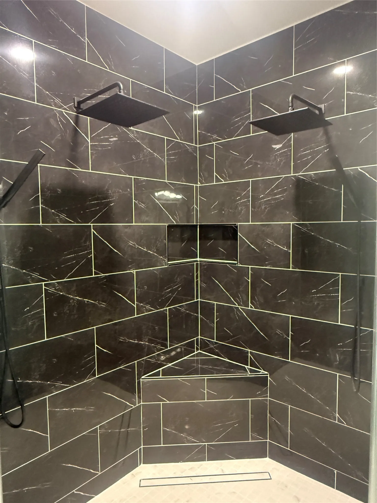 Shower enclosure featuring dark marbled-finish tile, dual matte black rain shower heads, a built-in recessed niche, a built-in corner shelf, and a linear drain