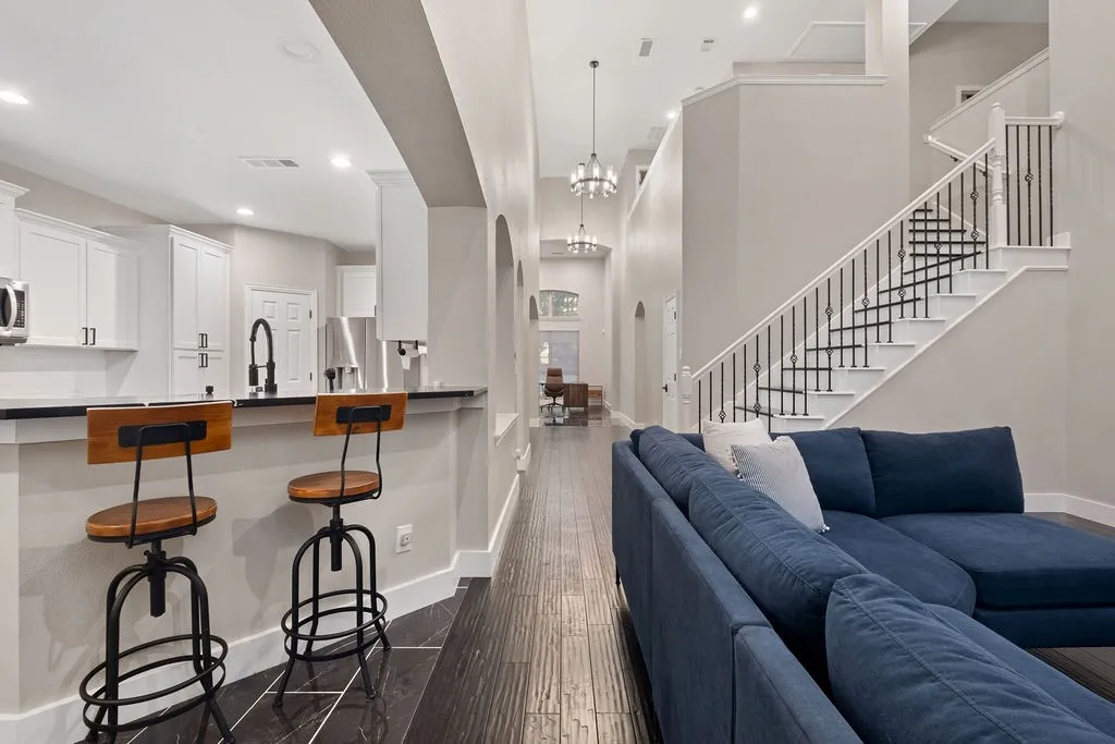 Open-concept living space with dark wood-finish flooring, a kitchen featuring white cabinetry and dark countertops, a breakfast bar, and a two-story entryway with a grand staircase and wrought iron balusters