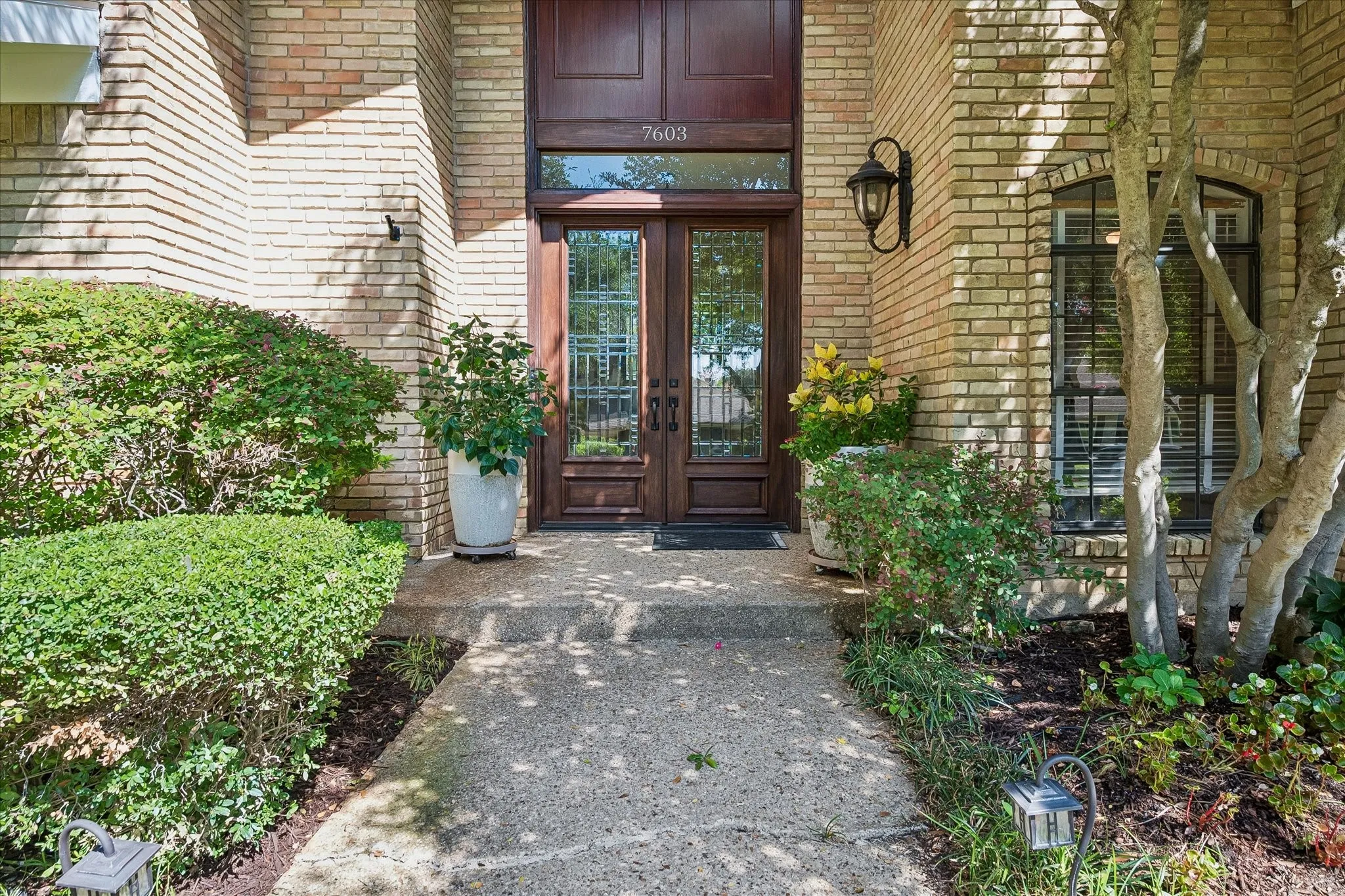 Brick exterior with a concrete walkway, double wood-finish entry doors, transom window, and an exterior wall lantern
