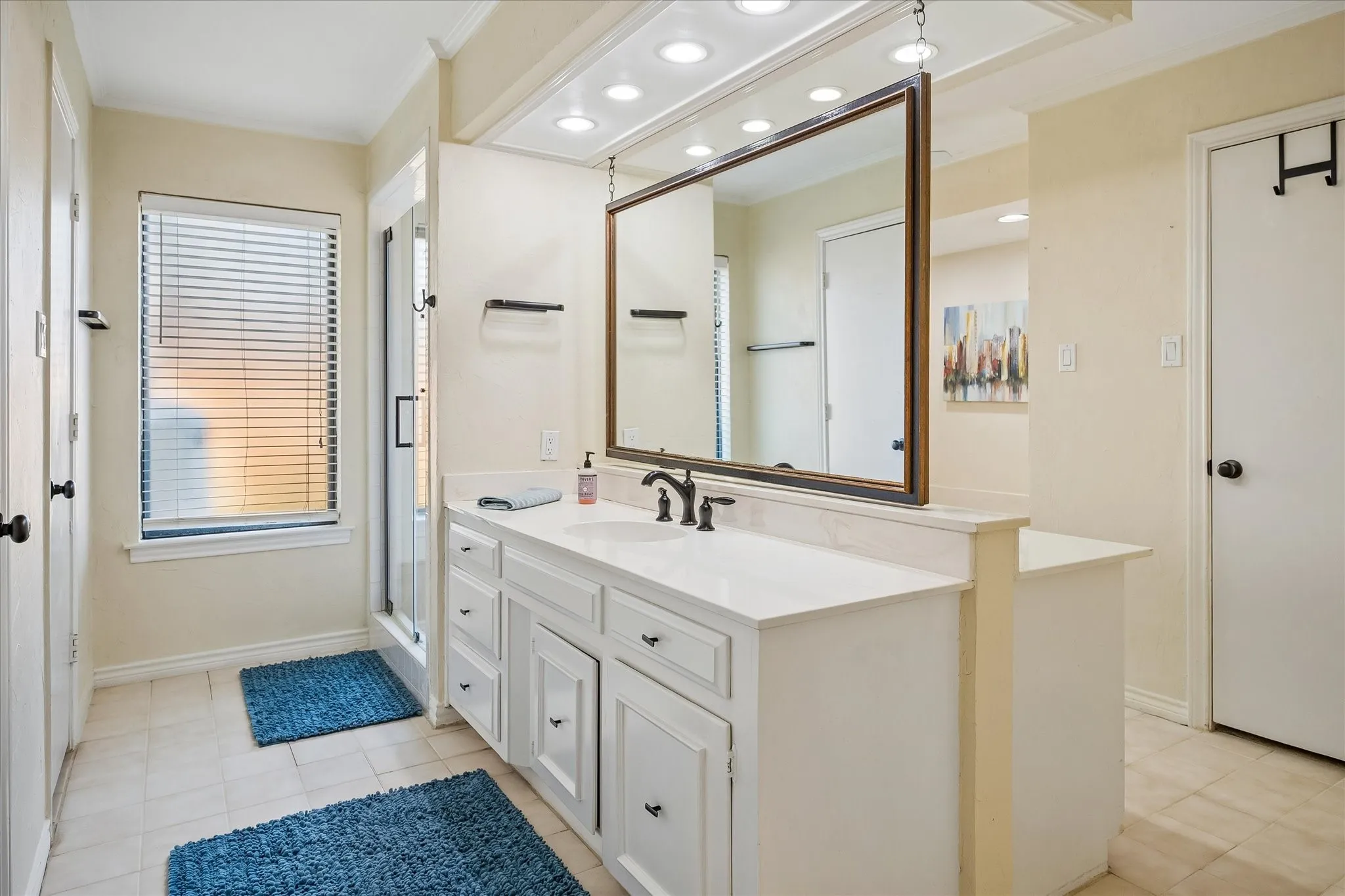 Spacious bathroom featuring a white vanity with integrated sink, large framed mirror, tiled flooring, and a glass-enclosed shower