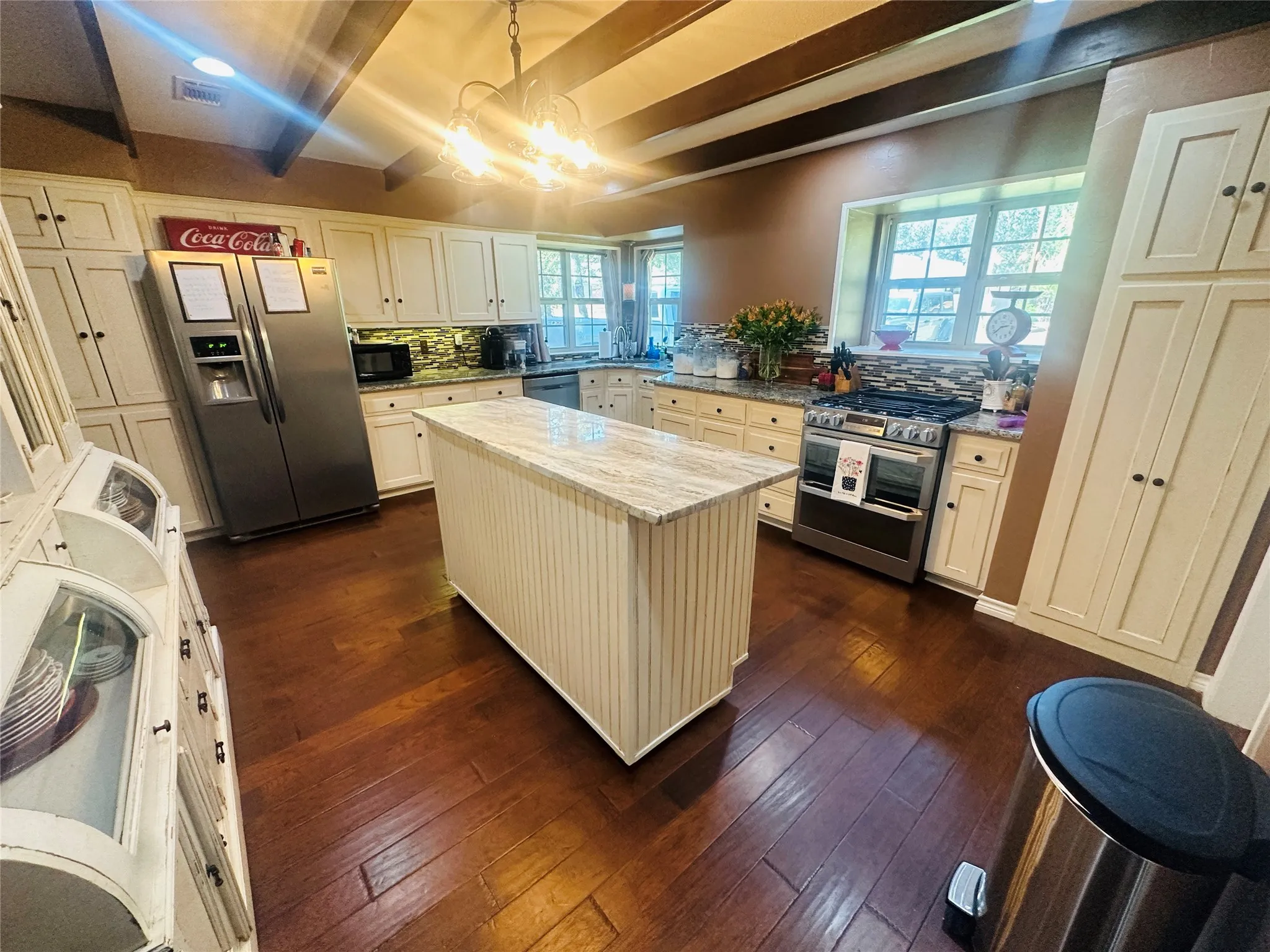 Kitchen featuring wood-finish flooring, an island with a light-toned countertop, white cabinetry, a tiled backsplash, and exposed ceiling beams