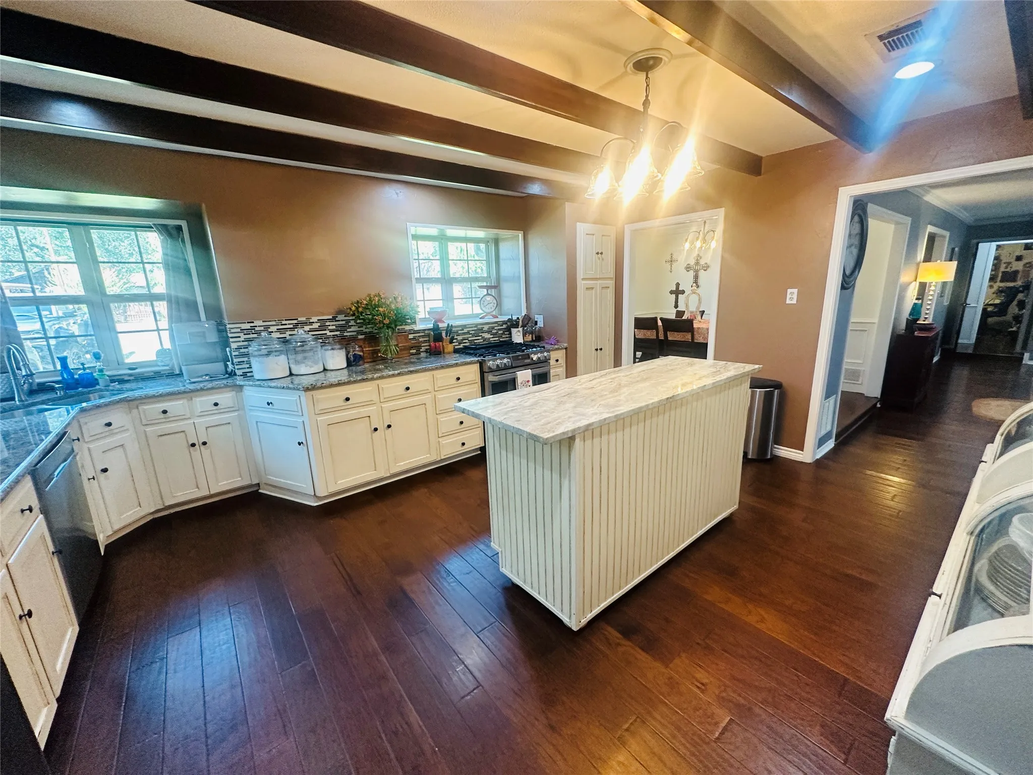 Kitchen featuring wood-finish flooring, white cabinetry, a center island with a light-toned countertop, a tile backsplash, and exposed ceiling beams