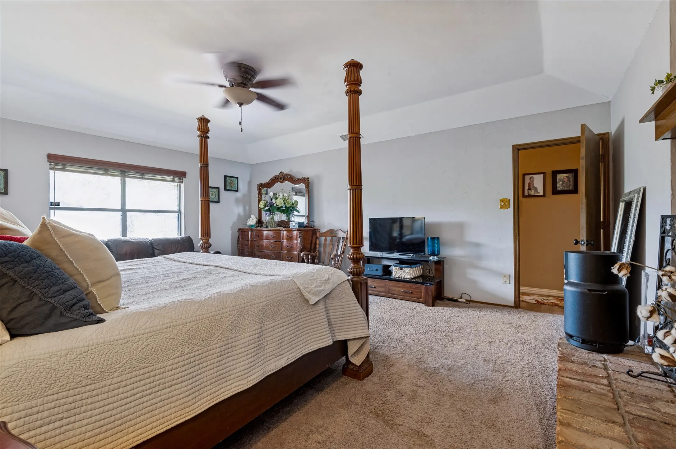 Spacious primary suite featuring a tray ceiling, ceiling fan, carpeted flooring and brick fireplace.