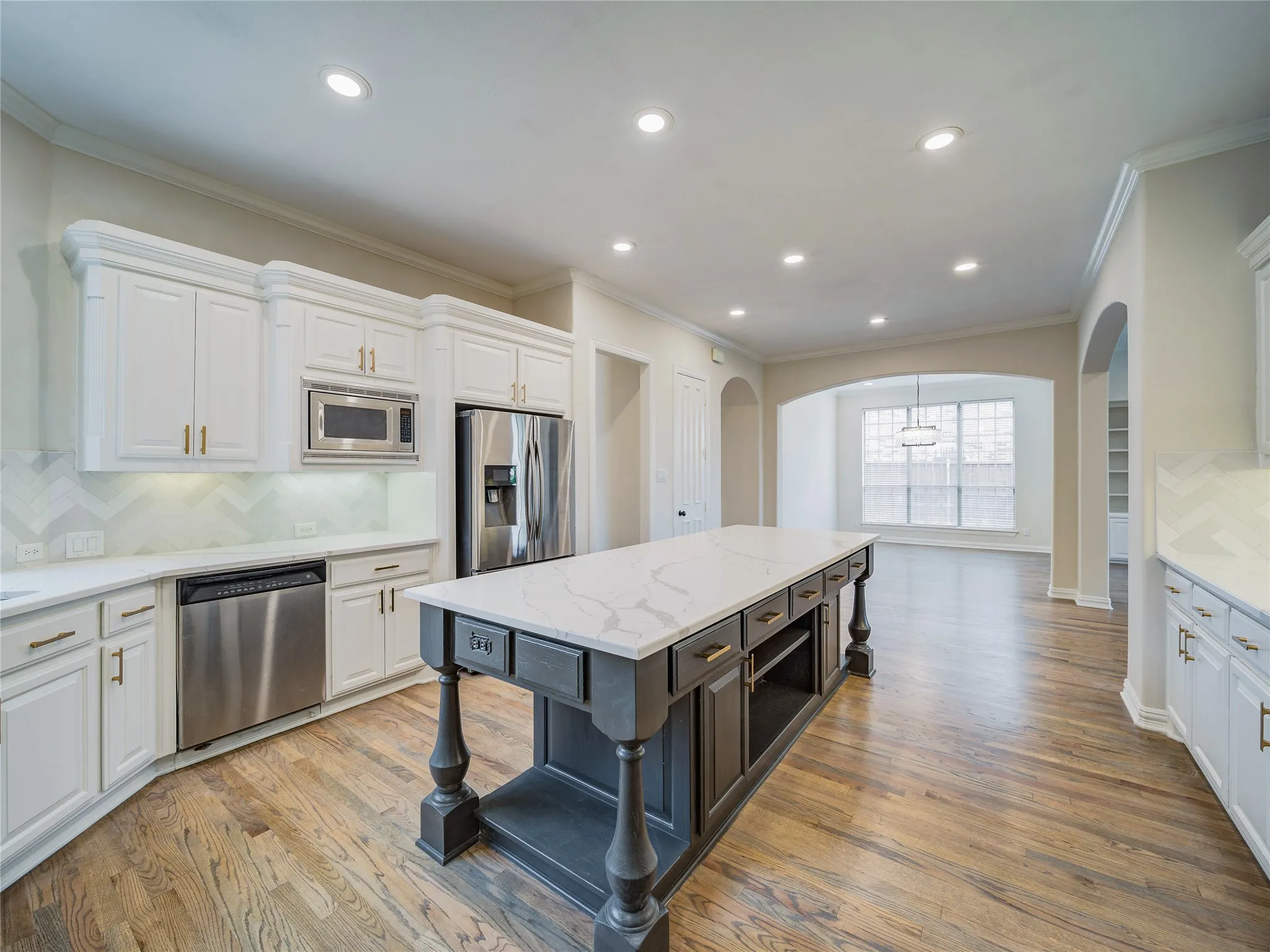 Kitchen featuring a large central island with a stone countertop, stainless steel appliances, white cabinetry with gold-tone hardware, a herringbone pattern backsplash, and wood-finish flooring