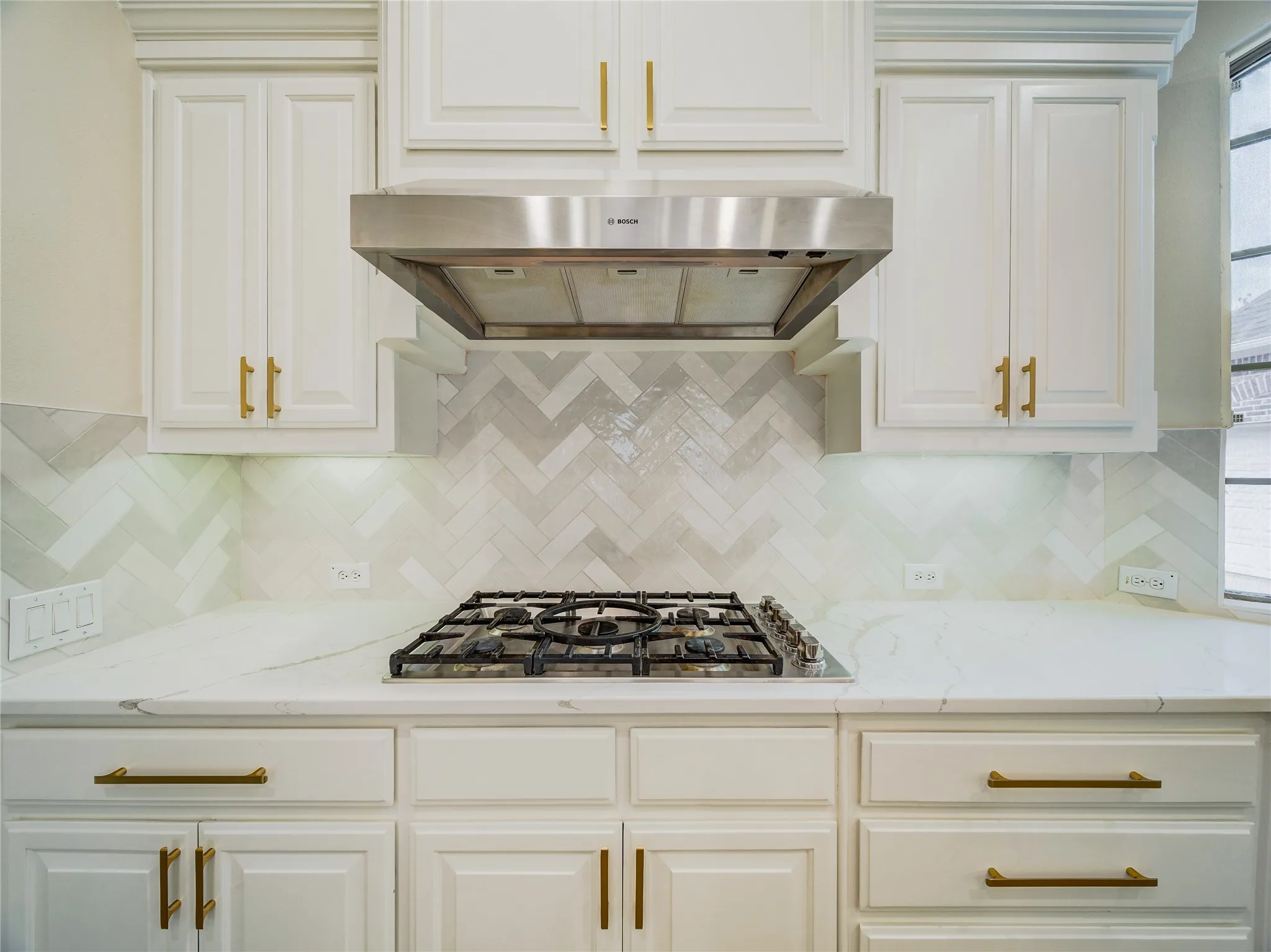 White cabinetry with gold-finish hardware, herringbone tile backsplash, stainless steel range hood, built-in gas cooktop, and light-toned countertops