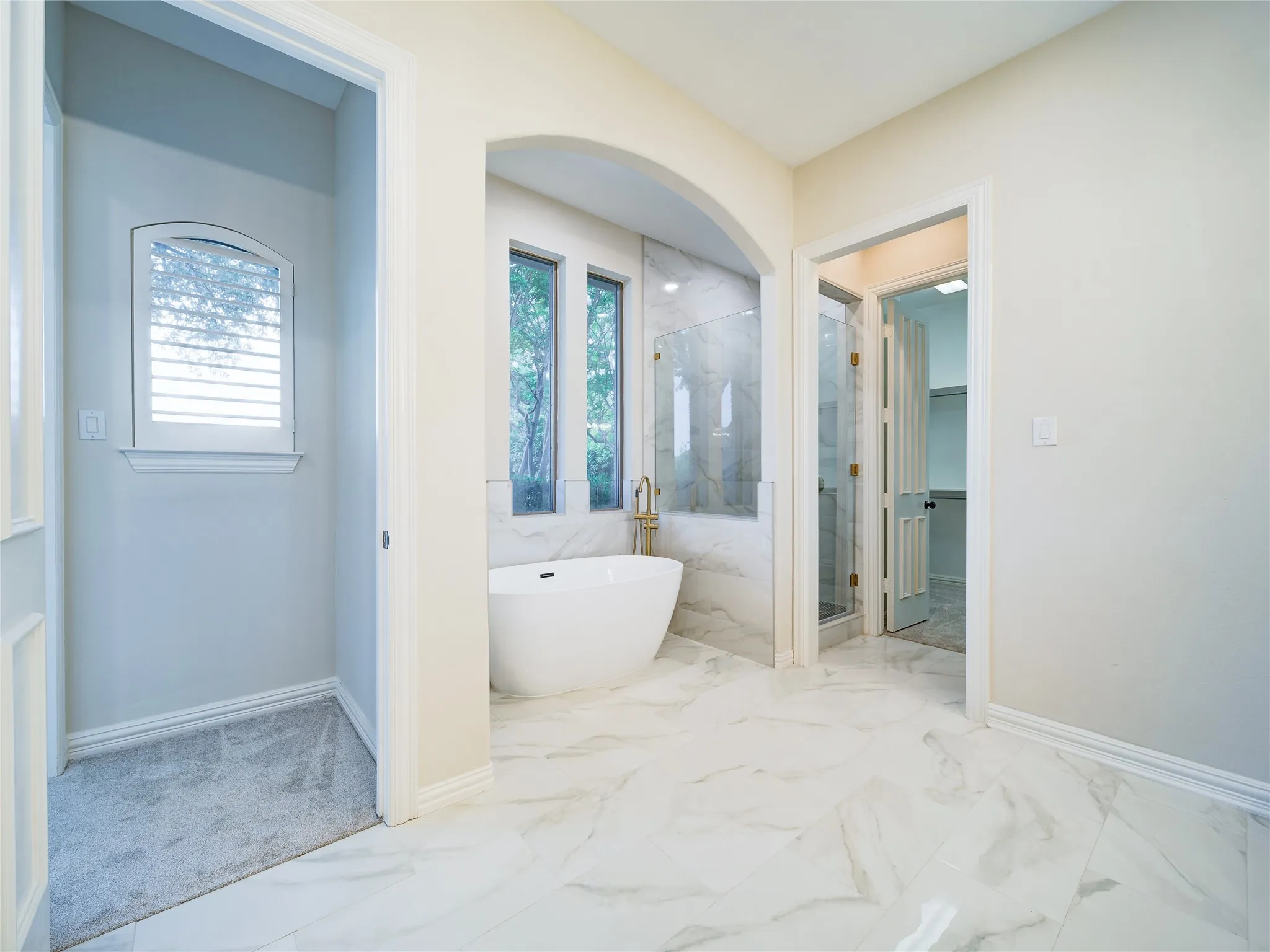 Freestanding bathtub set within an arched alcove, marble-look tiled shower enclosure with brass fixtures, large format marble-look floor tiles, and an arched window with plantation shutters