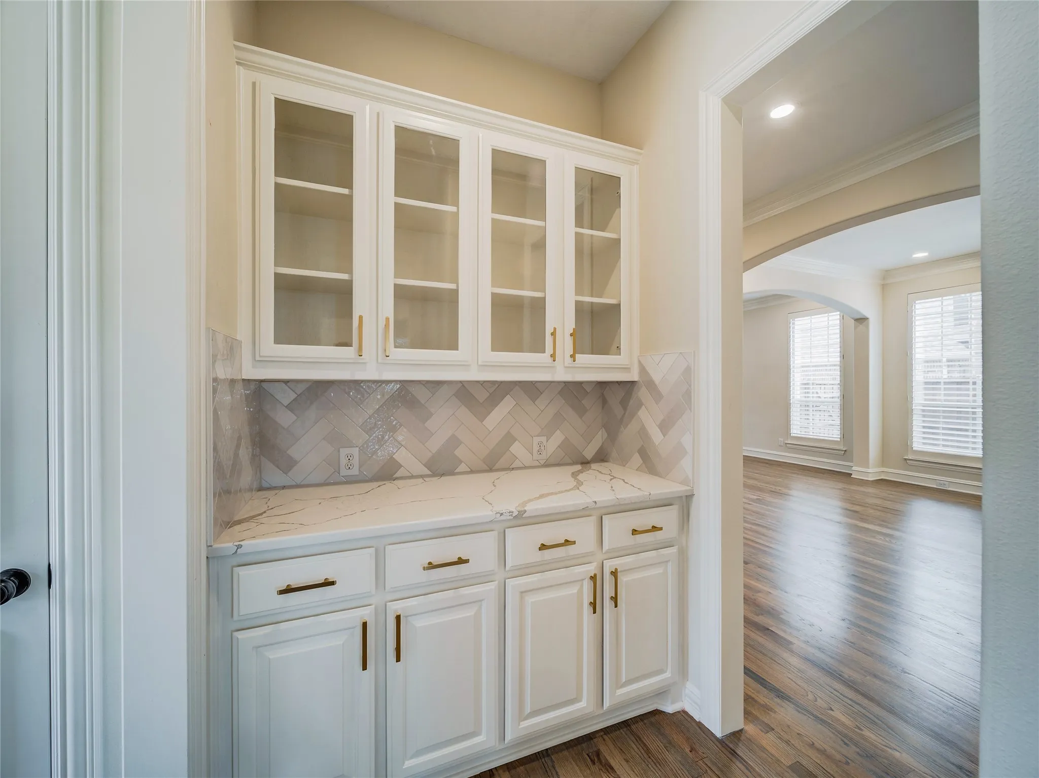 Elegant wet bar featuring cream-painted cabinetry with glass-front upper cabinets, brushed brass hardware, and a herringbone tile backsplash