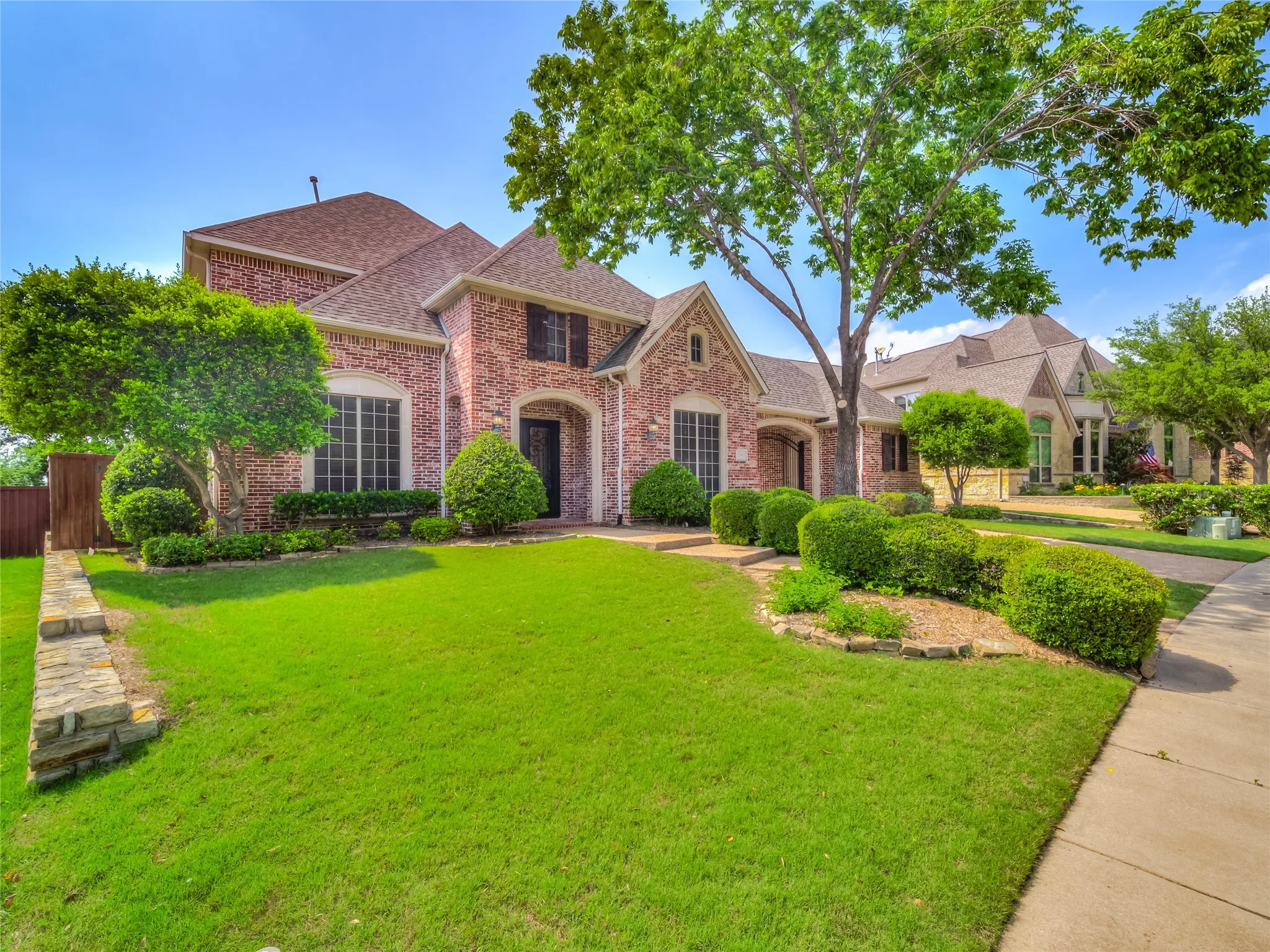 Elegant brick Colonial featuring a manicured front lawn, mature landscaping with boxwood hedges and specimen trees, concrete driveway, and multi-gabled roofline with architectural detailing