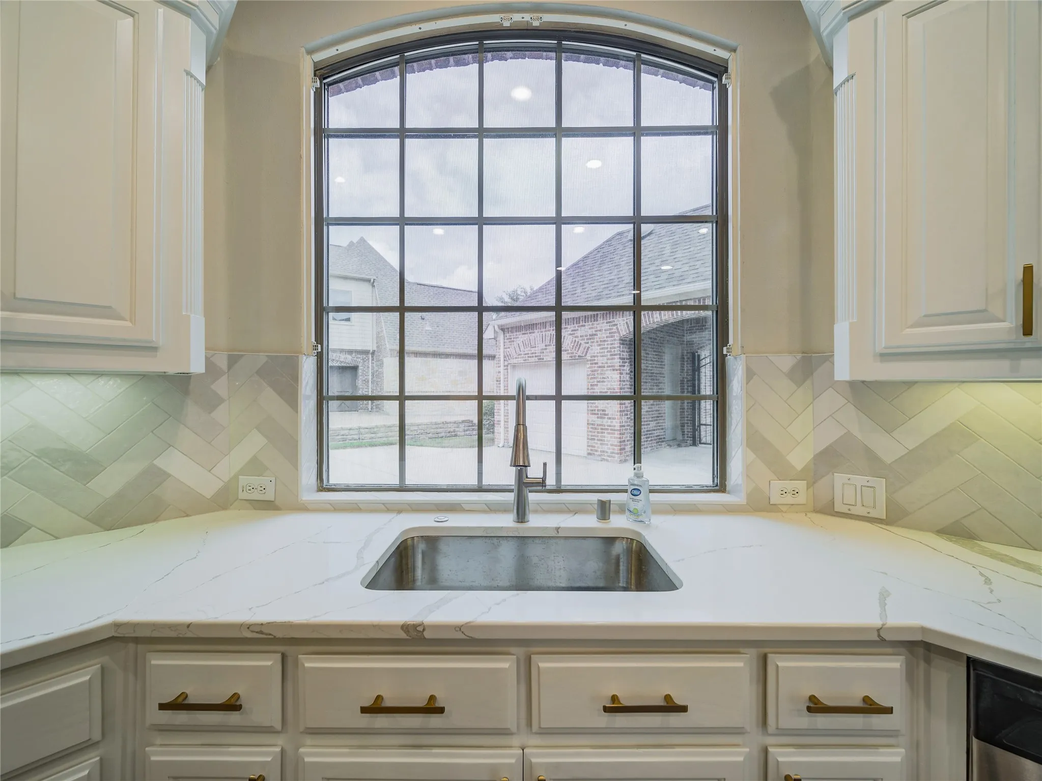 Chef's kitchen featuring a curved arched window with multiple panes, white cabinetry with brass hardware, white quartz countertops, and an undermount stainless steel sink with a polished chrome faucet