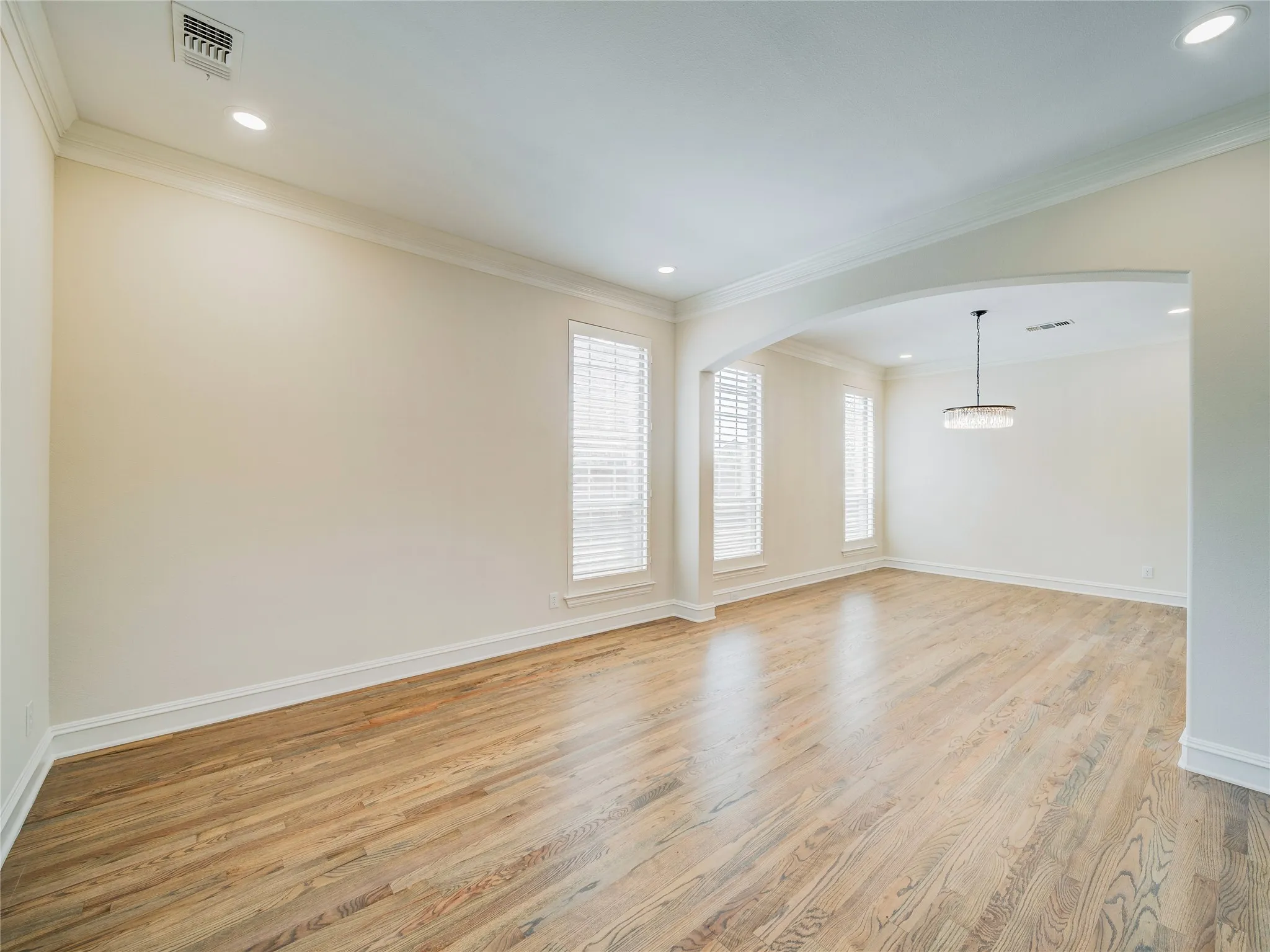 Hardwood flooring, recessed ceiling lighting, crown molding, multiple windows with blinds, and an arched doorway leading to an additional space with a suspended light fixture