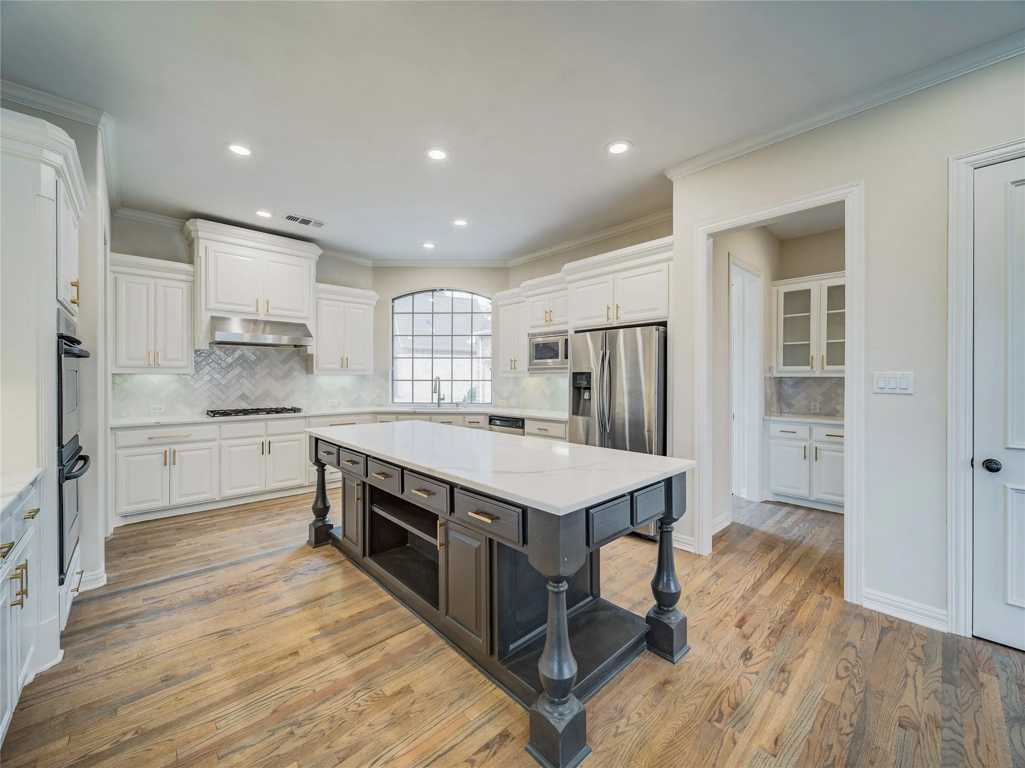 Gourmet kitchen featuring a central island with a light-toned countertop, white cabinetry with gold-finish hardware, stainless steel appliances, wood-finish flooring, and a decorative arched window