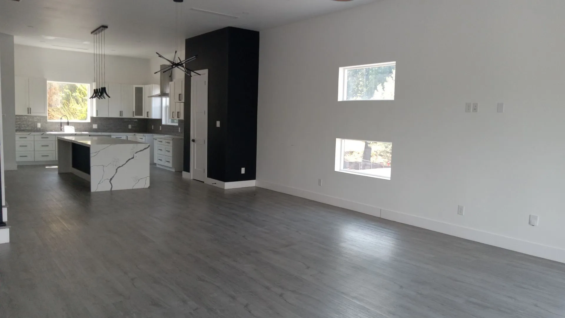 Open-concept interior featuring light wood-finish flooring, a contemporary kitchen with white cabinetry, a waterfall island, and a black accent wall