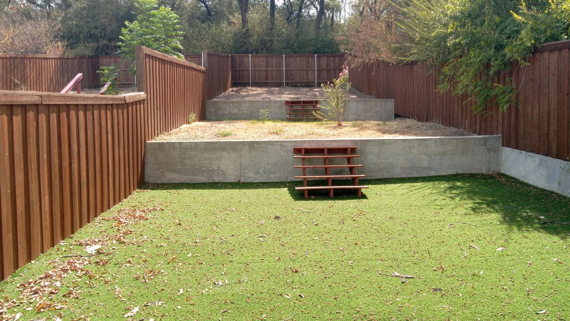 Tiered backyard with artificial turf, wood fencing, concrete retaining walls, and a small tree