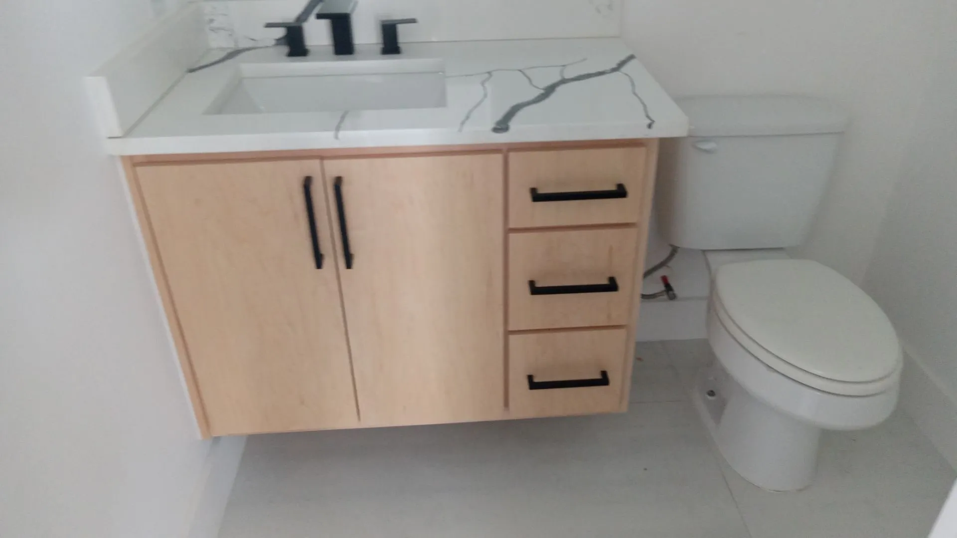 Light wood-finish vanity with black hardware, white countertop with gray veining, integrated rectangular sink, and matte black faucet