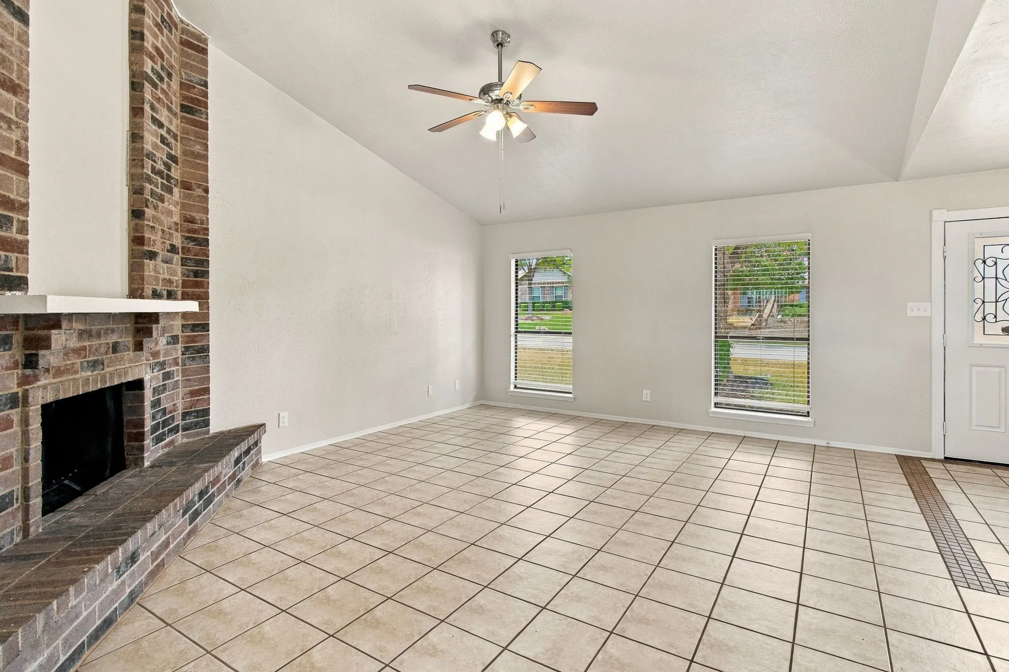 Brick fireplace with a white mantel, tiled hearth, vaulted ceiling, ceiling fan with light fixture, and tile flooring
