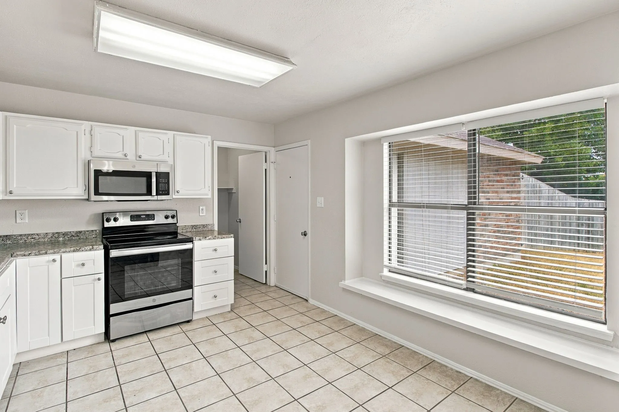 White cabinetry with chrome hardware, stainless steel appliances, granite countertops, tile flooring, and a large window with blinds
