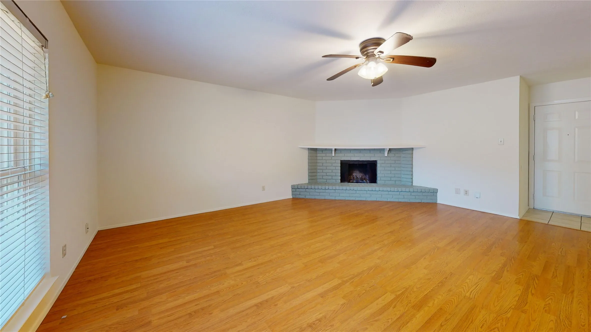 Hardwood-style flooring, brick fireplace with mantel, ceiling fan with light fixture, and window with blinds