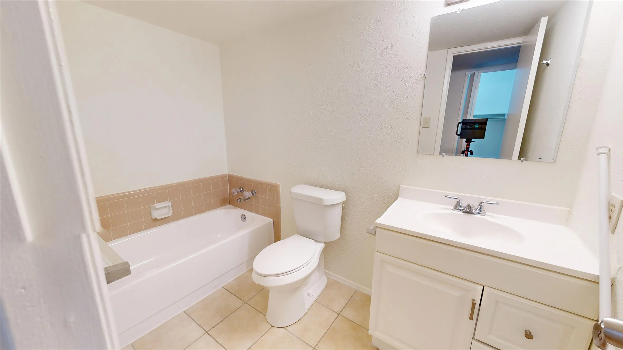 Ceramic tile flooring, white bathtub with tiled surround, white toilet, vanity with integrated sink, and wall-mounted mirror