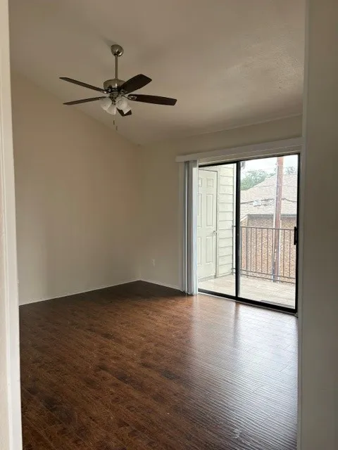 Wood-finish flooring, vaulted ceiling, ceiling fan with integrated lighting, and a sliding glass door providing access to an exterior balcony