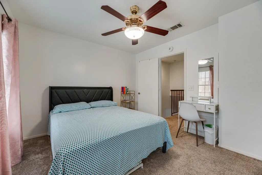 Textured headboard, ceiling fan with integrated lighting, carpeted flooring, and a mirrored vanity desk