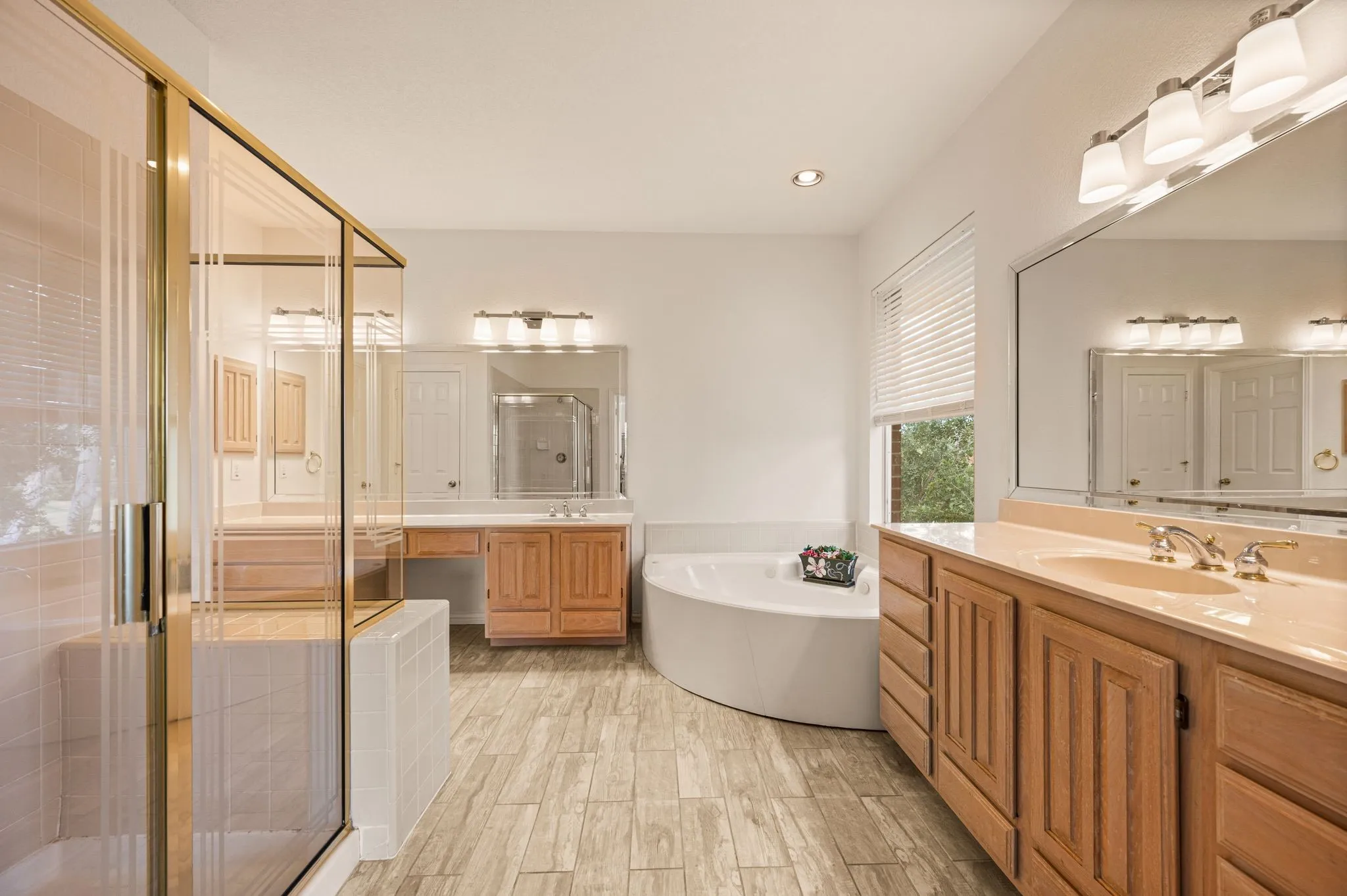 Spacious bathroom featuring a glass-enclosed shower, corner soaking tub, wood-finish vanity with dual sinks, and light wood-look flooring