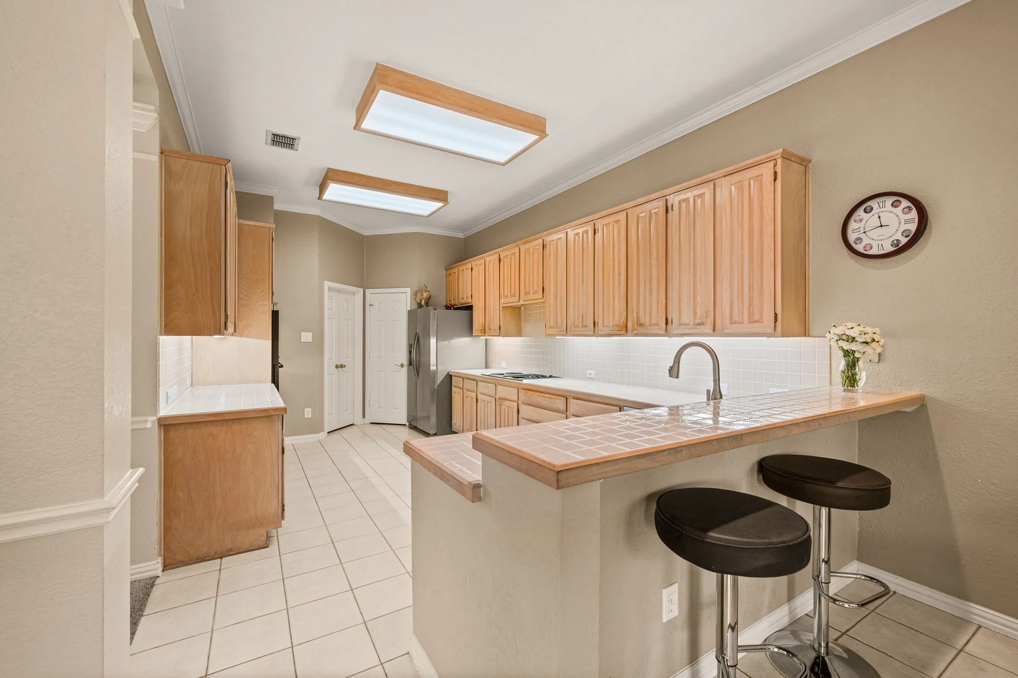 Kitchen featuring extensive wood cabinetry, white tiled flooring, a tiled backsplash, and an integrated breakfast bar with a stainless steel sink