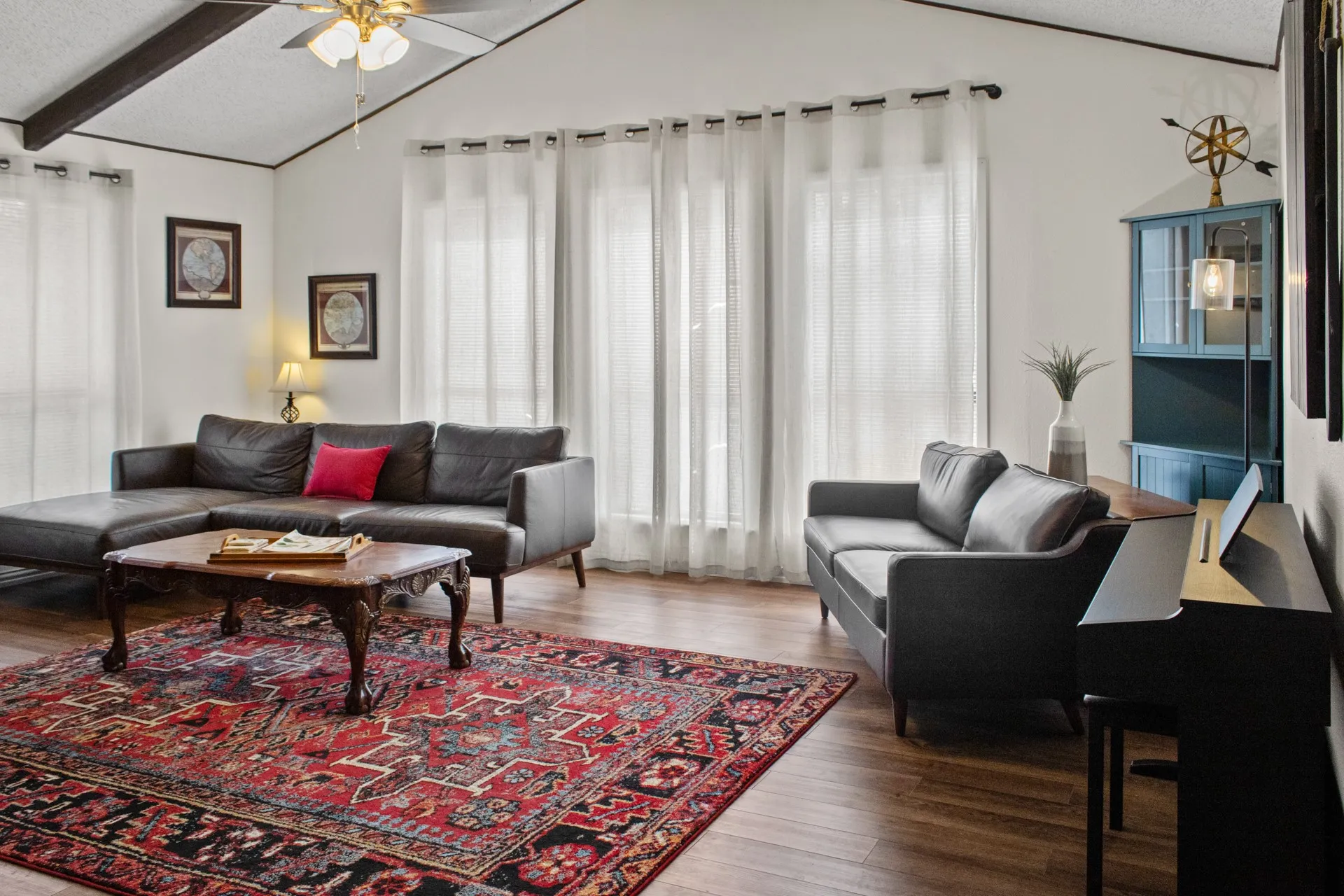 Vaulted ceiling with exposed beams, wood-finish flooring, and multiple large windows with sheer panel curtains