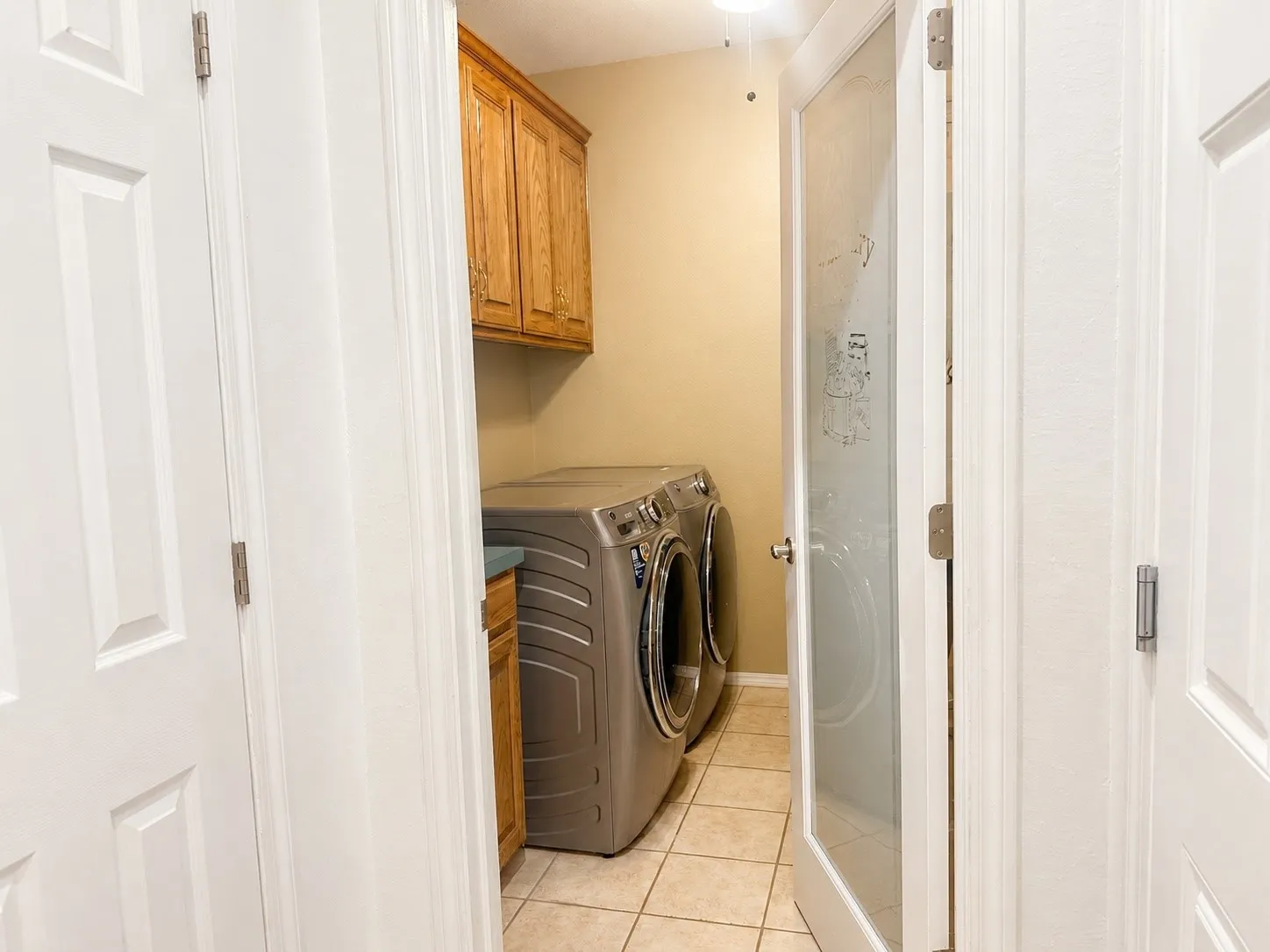 Dedicated laundry room featuring tile flooring, built-in cabinetry with wood-finish doors, a front-load washer and dryer unit, and bright overhead lighting for convenient home maintenance
