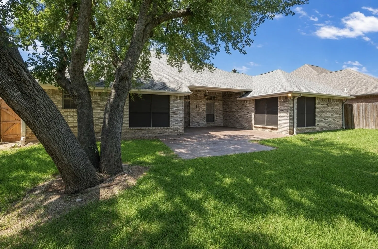 Brick exterior with a covered patio, paved hardscaping, mature trees, and a wooden privacy fence