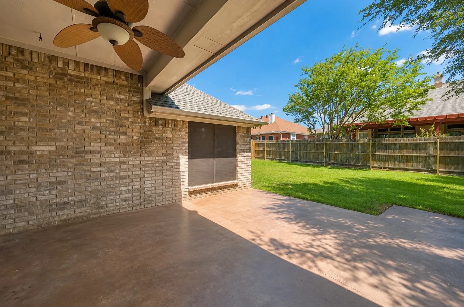 Expansive concrete patio with an attached ceiling fan, brick exterior wall, and fenced backyard with a manicured lawn