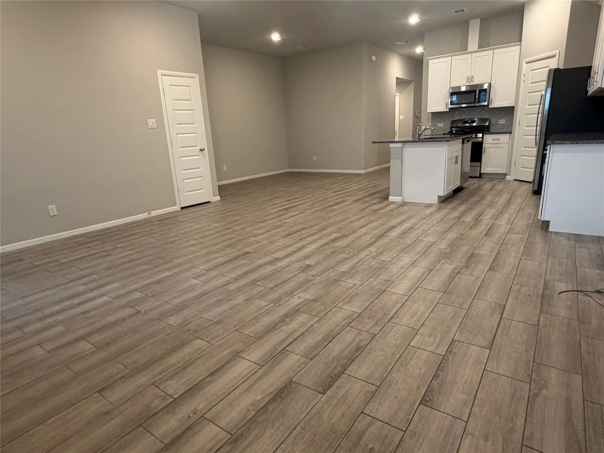 Open-concept living space featuring wood-finish tile flooring, recessed lighting, a kitchen island with a built-in sink, stainless steel appliances, and white cabinetry