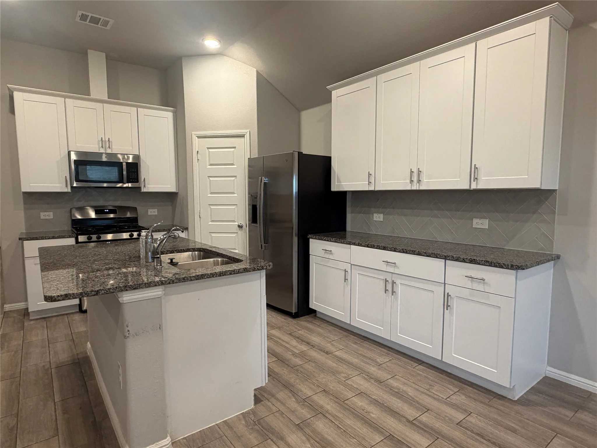 Kitchen featuring a central island with an undermount sink, granite countertops, white shaker-style cabinetry, stainless steel appliances, and wood-finish flooring