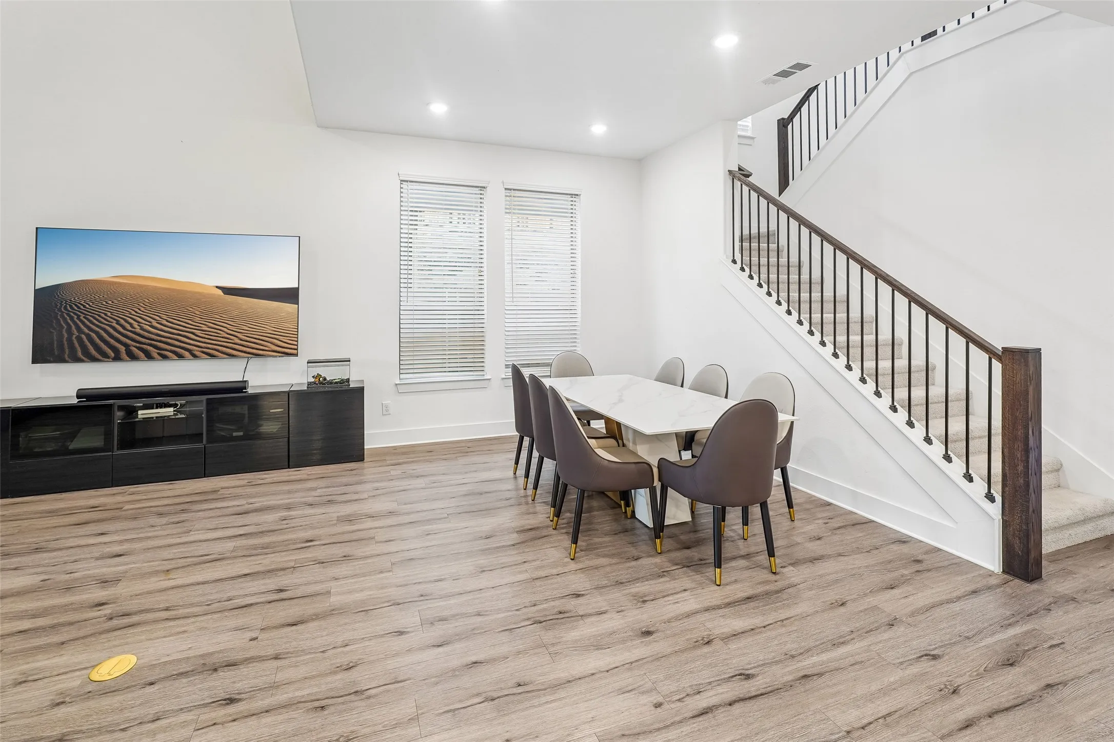 Vaulted ceiling, recessed lighting, wood-finish flooring, two windows with blinds, and a staircase with a dark wood handrail and black balusters