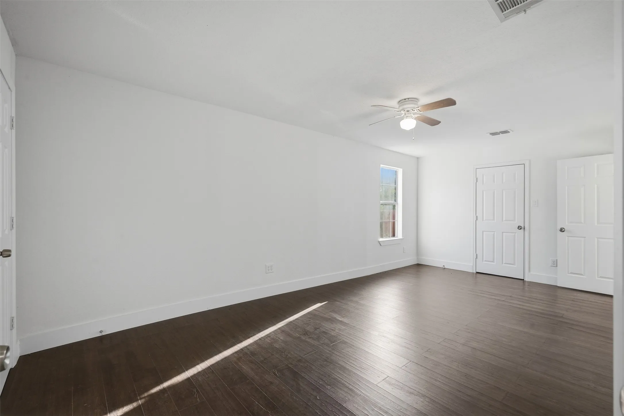 Spacious room featuring wood-finish flooring, a ceiling fan with integrated lighting, a window, and two paneled doors