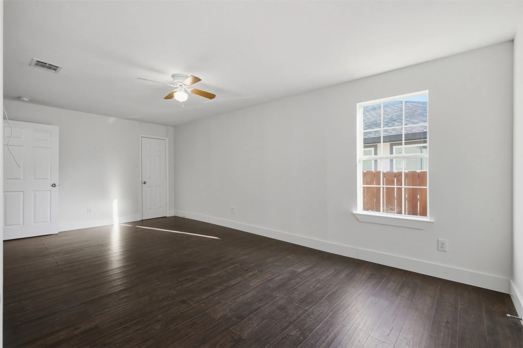 Spacious room featuring dark wood-finish flooring, a white window frame, and a ceiling fan with light fixture