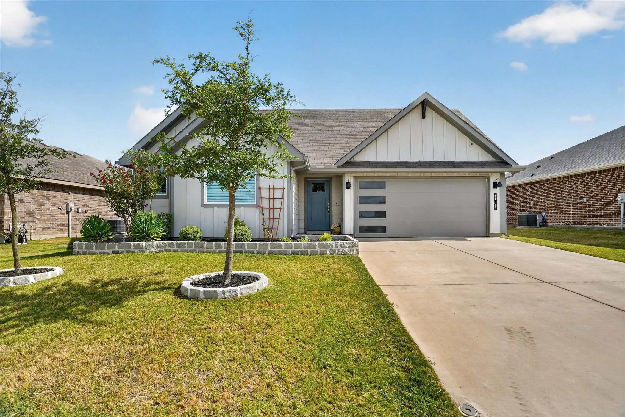 Contemporary single-story residence with a modern garage door, concrete driveway, and landscaped front yard featuring mature trees and a decorative stone planter