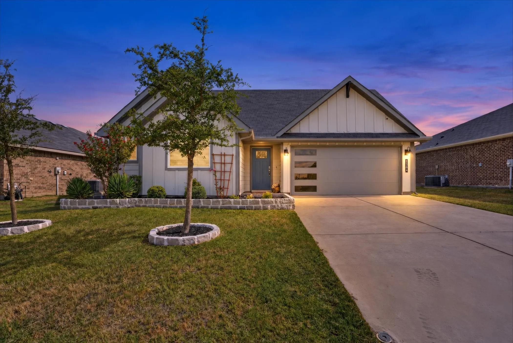Modern single-story residence with a concrete driveway and attached garage featuring contemporary glass panels, accent lighting as well as stacked stone flowerbed.