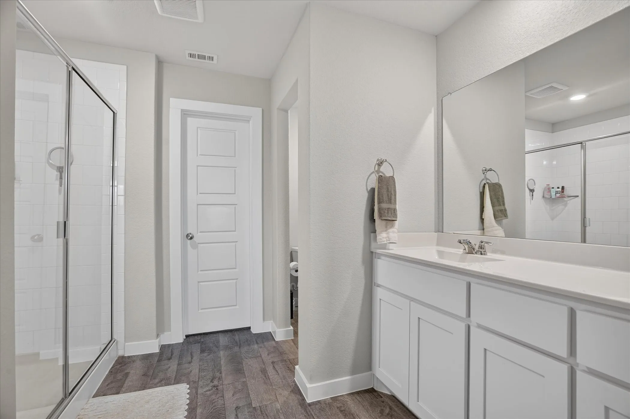 Spacious bathroom featuring a glass-enclosed shower, wood-finish flooring, a white vanity with integrated sink, and a large wall-mounted mirror