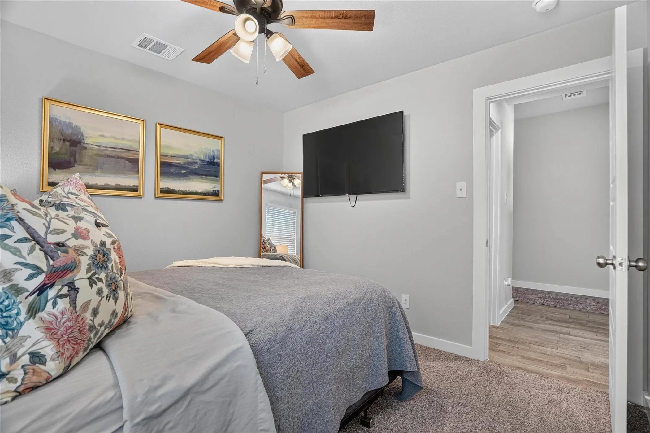 Carpeted flooring, ceiling fan with light fixture, wall-mounted television, and a doorway leading to a space with wood-finish flooring
