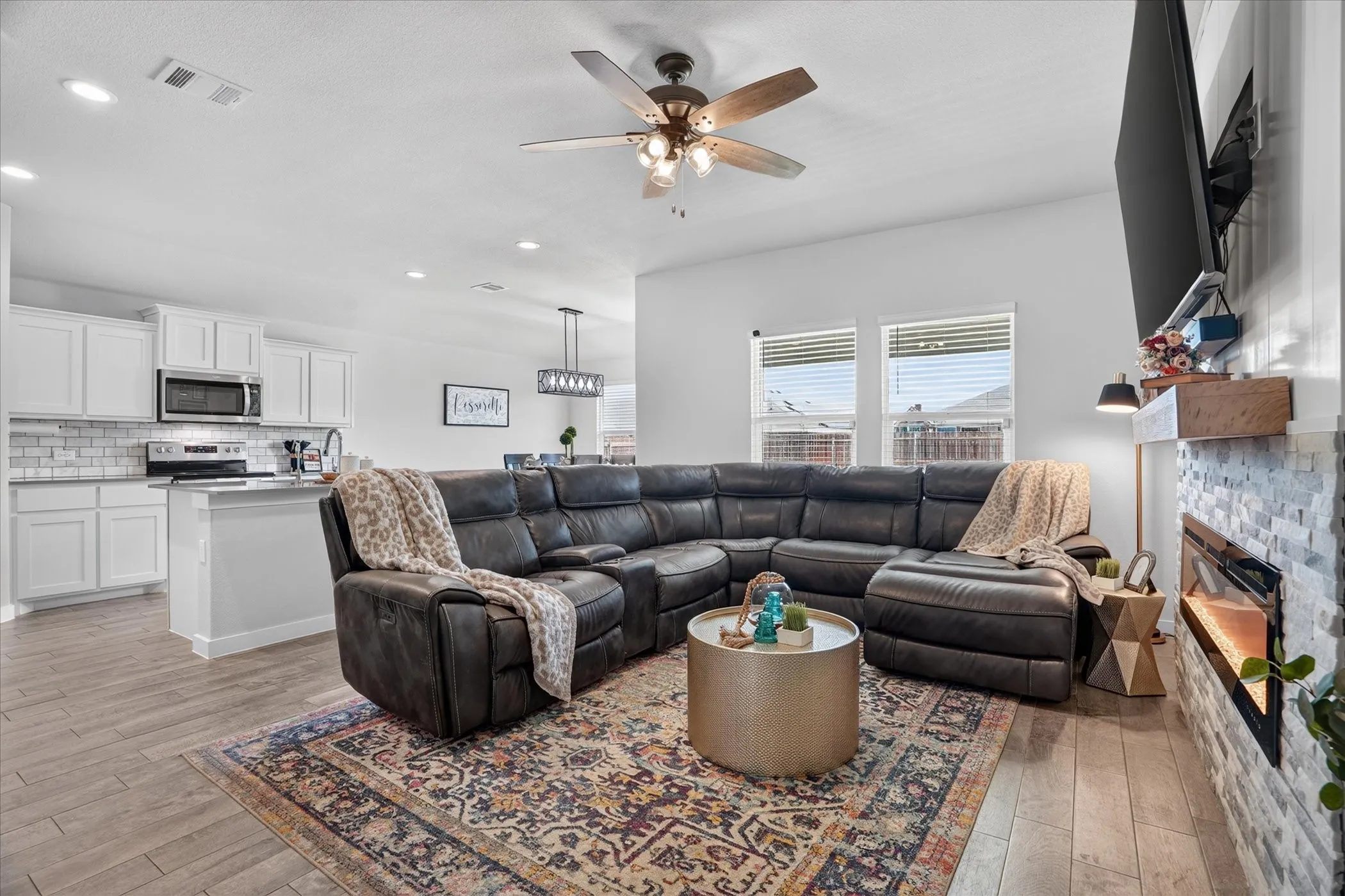 Open-concept living area featuring wood-finish flooring, a ceiling fan, and an electric fireplace with a stacked stone surround and rustic wood mantel