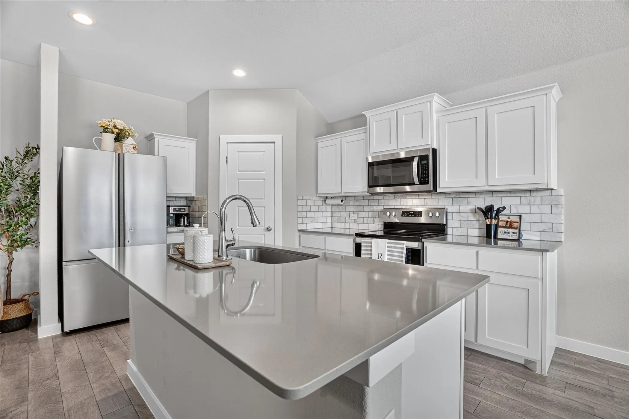 Kitchen featuring an island with an undermount sink, stainless steel appliances, white cabinetry, a subway tile backsplash, and wood-finish flooring