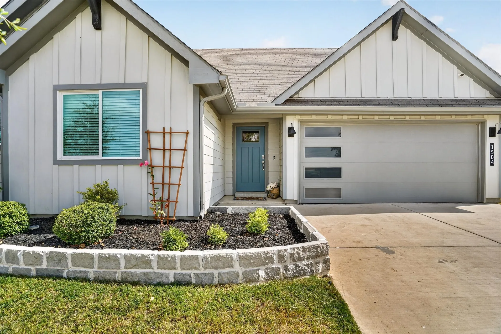 Board and batten siding with contrasting trim accents, stone-faced planter with established landscaping, and a contemporary garage door featuring horizontal window inserts