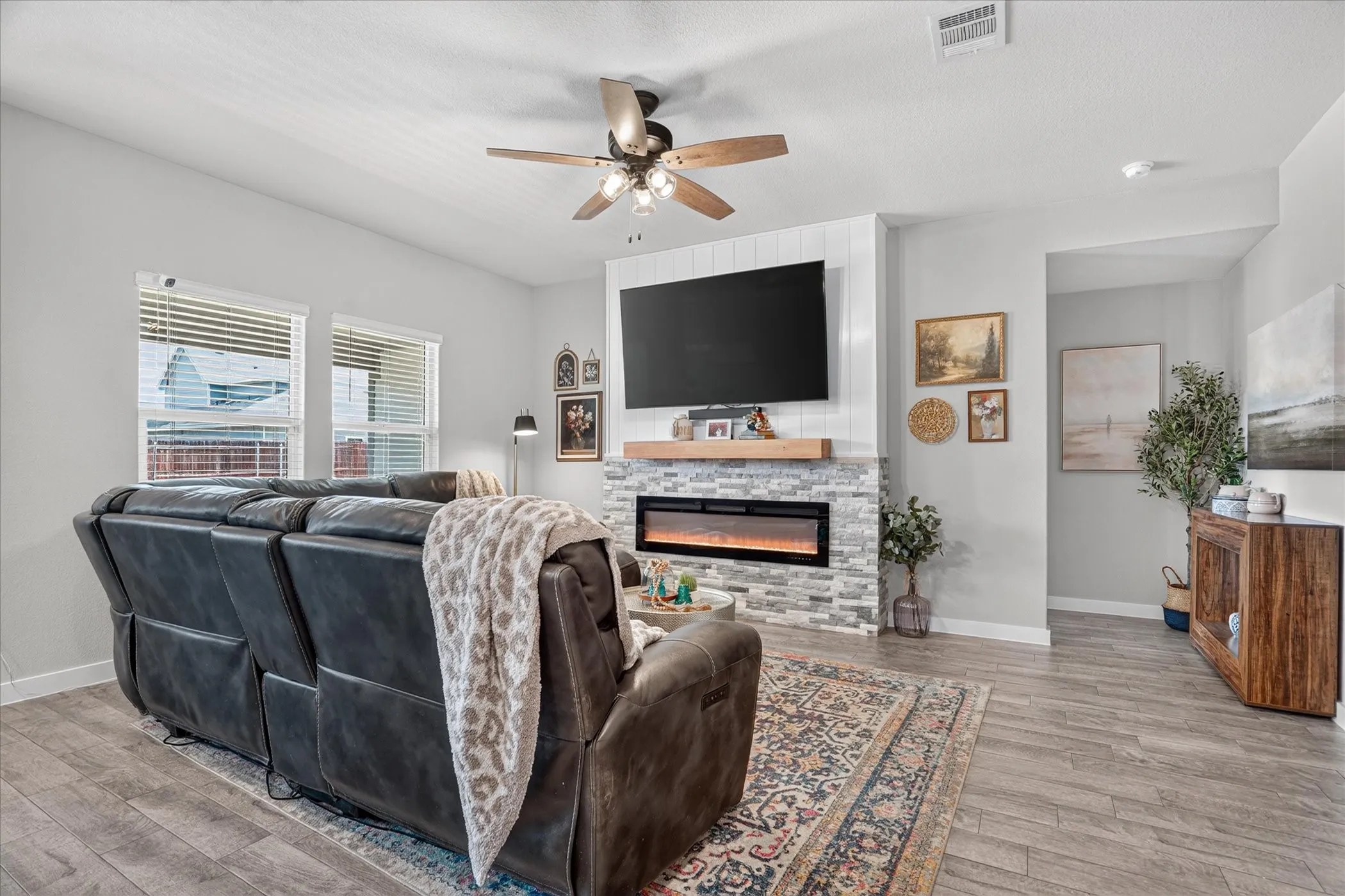 Living area featuring wood-finish flooring, a stone-clad fireplace with a wood mantel, a ceiling fan with integrated lighting, and multiple windows